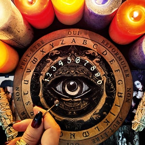May include: A wooden Ouija board with an all-seeing eye design, surrounded by lit candles. The board features the letters of the alphabet, numbers 1-9 and 0, and the words "Oui", "Peut-&ecirc;tre", "Non", and "Reformuler".