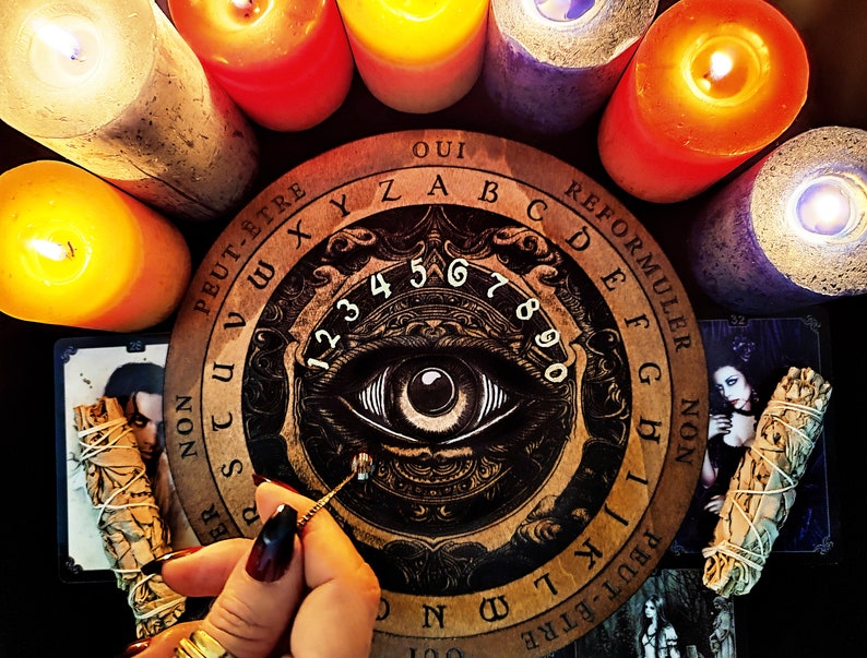 May include: A wooden Ouija board with an all-seeing eye design, surrounded by lit candles. The board features the letters of the alphabet, numbers 1-9 and 0, and the words "Oui", "Peut-&ecirc;tre", "Non", and "Reformuler".