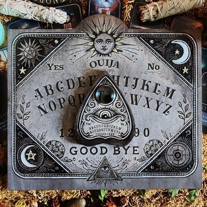 May include: A gray Ouija board with a planchette, featuring the alphabet, numbers, and the words "Yes," "No," and "Good Bye." The board has a sun and moon design, surrounded by tarot cards and candles.