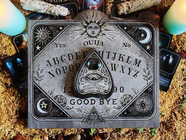 May include: A gray Ouija board with a planchette, featuring the alphabet, numbers, and the words "Yes," "No," and "Good Bye." The board has a sun and moon design, surrounded by tarot cards and candles.