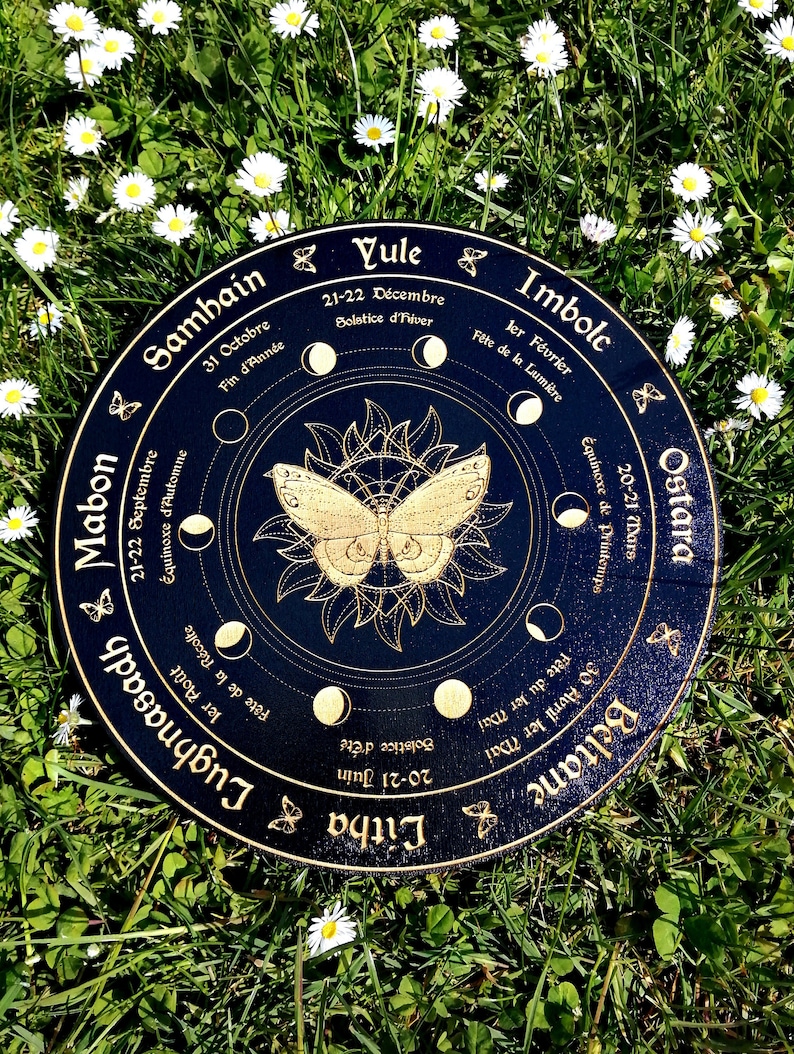 May include: A black wooden wheel with gold accents depicting the eight sabbats of the Wheel of the Year. The wheel features a butterfly design in the center and the names of each sabbat in French and English. The wheel is surrounded by a ring of white daisies.