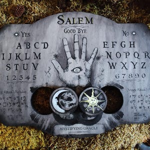 K&ouml;nnte beinhalten: Ein Ouija-Brett aus Holz mit einem schwarz-wei&szlig;en Design. Das Brett zeigt die Buchstaben des Alphabets, Zahlen und die W&ouml;rter "Ja", "Nein", "Leb wohl" und "Salem". Das Brett hat auch eine Hand mit einem Auge in der Mitte. Das Brett ist von Moos umgeben.