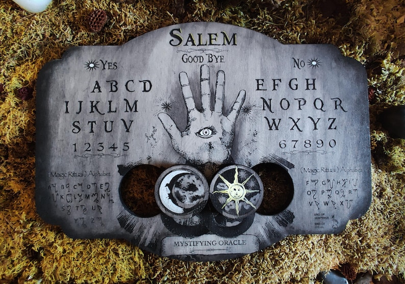 K&ouml;nnte beinhalten: Ein Ouija-Brett aus Holz mit einem schwarz-wei&szlig;en Design. Das Brett zeigt die Buchstaben des Alphabets, Zahlen und die W&ouml;rter "Ja", "Nein", "Leb wohl" und "Salem". Das Brett hat auch eine Hand mit einem Auge in der Mitte. Das Brett ist von Moos umgeben.
