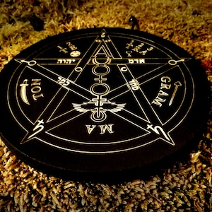 May include: A black circular wooden board with a gold pentagram design. The pentagram features the words "GRAM", "TON", "TIB", "NOL", and "MA" around the points. The centre of the pentagram has a winged symbol with a circle and a cross.