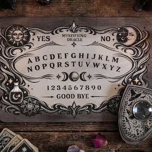 May include: A rectangular wooden spirit board featuring the text Mystifying Oracle, the alphabet, numbers zero through nine, and the words Yes, No, and Good Bye. The board includes illustrations of a sun, moon, stars, and a potion bottle. A heart-shaped planchette with a central lens and occult symbols rests on the board.