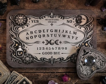 May include: A rectangular wooden spirit board featuring the text Mystifying Oracle, the alphabet, numbers zero through nine, and the words Yes, No, and Good Bye. The board includes illustrations of a sun, moon, stars, and a potion bottle. A heart-shaped planchette with a central lens and occult symbols rests on the board.