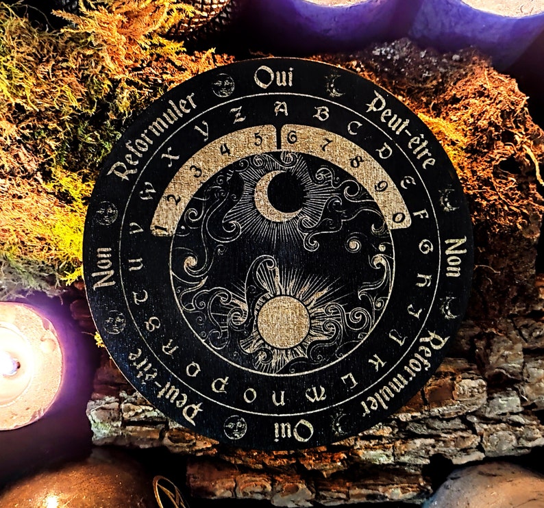 May include: A black wooden Ouija board with gold lettering and symbols. The board features a crescent moon and sun design in the centre. The text on the board reads "Oui", "Peut-&ecirc;tre", "Non", and "Reformuler".