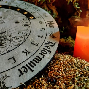 May include: A wooden planchette board with sun and moon illustrations and the word "Reformuler" is next to a lit orange candle. The board has letters and numbers around the edge. The scene is set on a bed of moss.