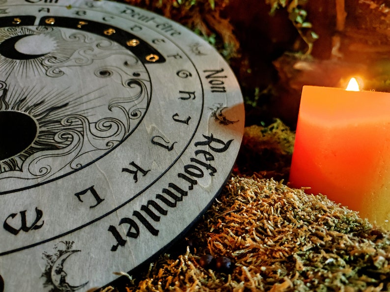 May include: A wooden planchette board with sun and moon illustrations and the word "Reformuler" is next to a lit orange candle. The board has letters and numbers around the edge. The scene is set on a bed of moss.