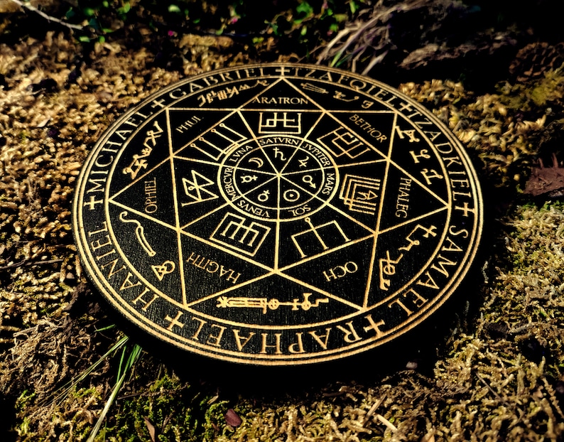 K&ouml;nnte beinhalten: Ein schwarzer Holzkreis mit goldenen Buchstaben und Symbolen. Der Kreis zeigt die Namen von Engeln und Planeten. Der Kreis ist von einem Ring aus goldenen Buchstaben umgeben.