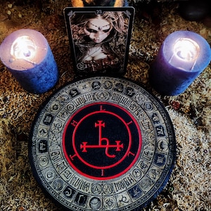 May include: A black and red sigil with a cross and a hook, surrounded by a circle of symbols, on a black and gold background. The sigil is on a round wooden board. Two black candles and a tarot card are in the background.