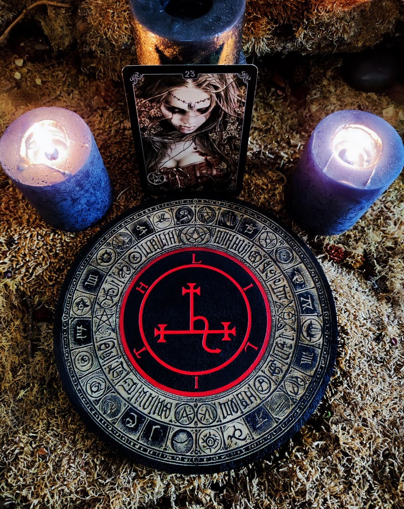 May include: A black and red sigil with a cross and a hook, surrounded by a circle of symbols, on a black and gold background. The sigil is on a round wooden board. Two black candles and a tarot card are in the background.
