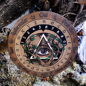 May include: A wooden Ouija board with a black and white illustration of an eye inside a triangle surrounded by green leaves. The board has numbers 1-9, letters A-Z, and the words "Oui", "Peut-&ecirc;tre", "Non", and "Reformuler" written around the edge.