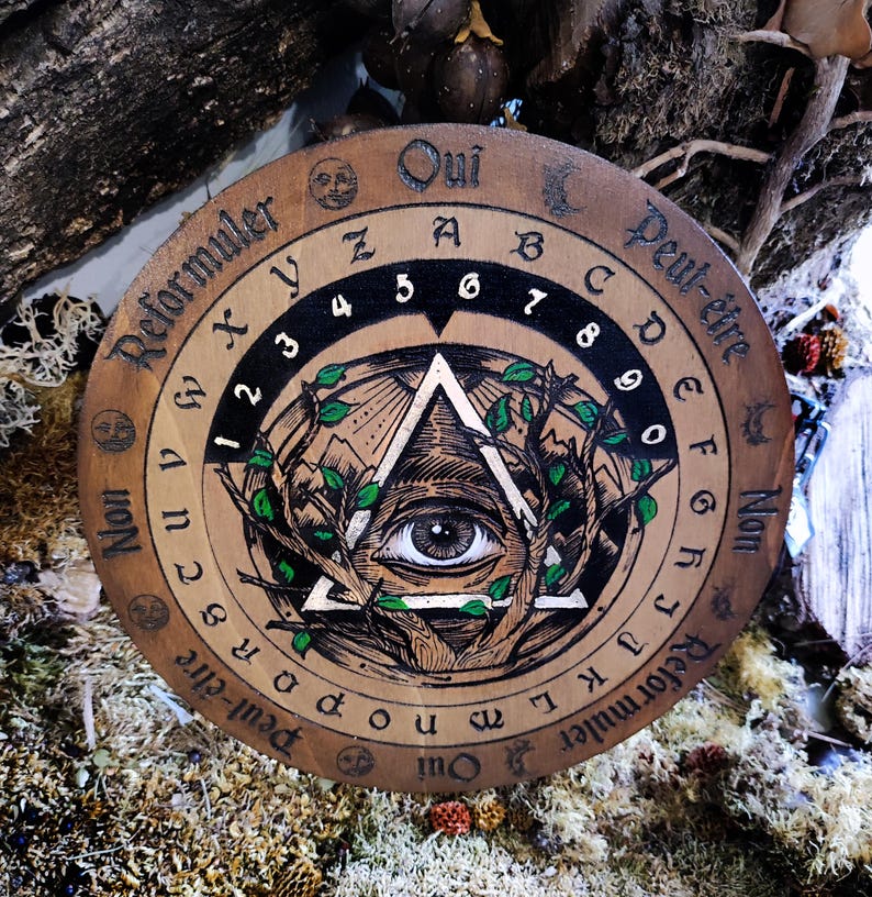 May include: A wooden Ouija board with a black and white illustration of an eye inside a triangle surrounded by green leaves. The board has numbers 1-9, letters A-Z, and the words "Oui", "Peut-&ecirc;tre", "Non", and "Reformuler" written around the edge.