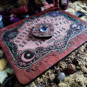 May include: A wooden Ouija board with a planchette in the center. The board has a dark red stain and features a decorative design with green, black, and gold accents. The board includes the alphabet, numbers 1-9 and 0, and the words "yes", "no", "good bye", and "Ouija".