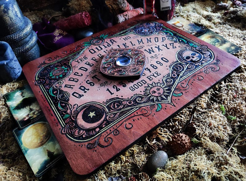 May include: A wooden Ouija board with a planchette in the center. The board has a dark red stain and features a decorative design with green, black, and gold accents. The board includes the alphabet, numbers 1-9 and 0, and the words "yes", "no", "good bye", and "Ouija".