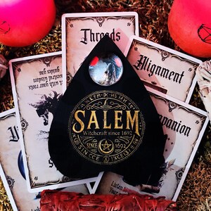 May include: A black teardrop-shaped pendant with a gold inscription that reads "SALEM Witchcraft since 1692". The pendant has a small circular glass window with a red and black image inside.