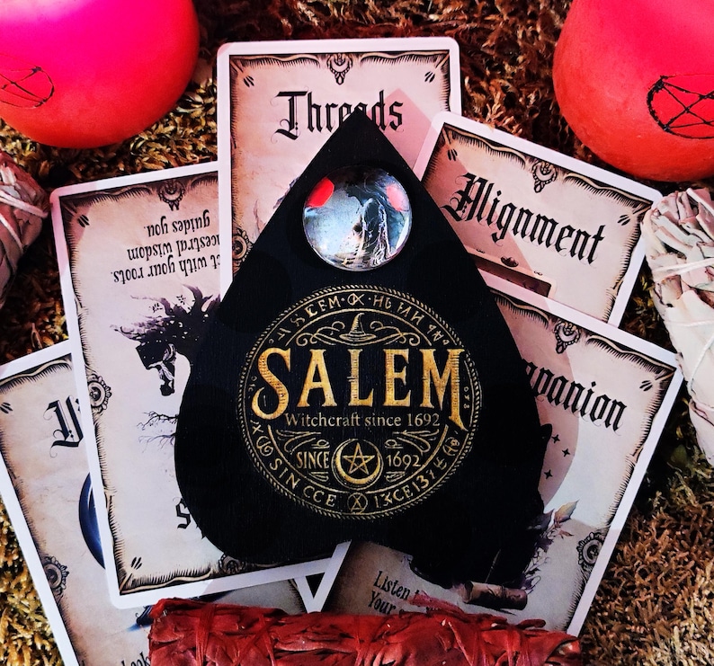 May include: A black teardrop-shaped pendant with a gold inscription that reads "SALEM Witchcraft since 1692". The pendant has a small circular glass window with a red and black image inside.