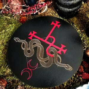 May include: A black circular coaster with a red cross and a gold snake design. The snake is intertwined with the cross and the design is on a black background.