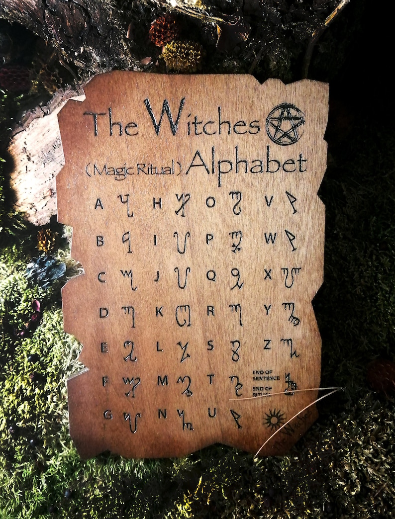 Puede incluir: Una placa de madera con fondo marr&oacute;n y texto negro. El texto dice "The Witches (Magic Ritual) Alphabet" e incluye un s&iacute;mbolo de pentagrama. La placa presenta una cuadr&iacute;cula de letras con s&iacute;mbolos correspondientes.