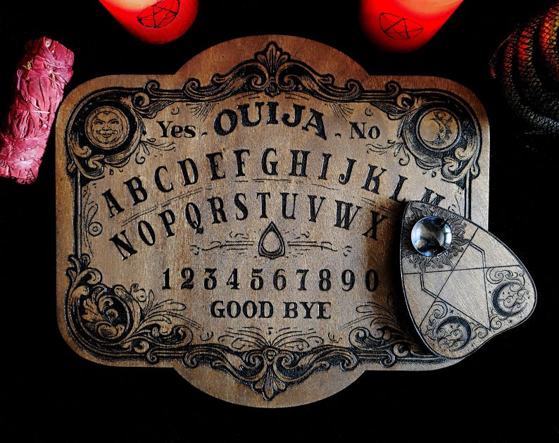 May include: A vintage-style Ouija board made of wood, featuring engraved letters, numbers, and the words "Yes," "No," and "Goodbye." The planchette has a clear gem. The board is surrounded by candles and a bundle of sage.