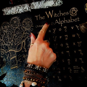 May include: A black background with a gold tree of life design and a gold witches alphabet. The alphabet is written in a stylized font and includes symbols for each letter. The text "The Witches Alphabet" is written at the top of the image.