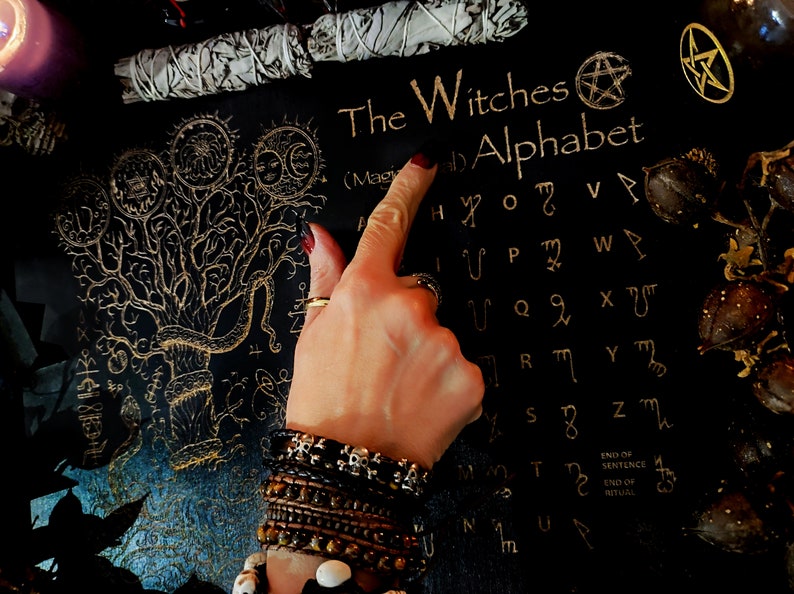 May include: A black background with a gold tree of life design and a gold witches alphabet. The alphabet is written in a stylized font and includes symbols for each letter. The text "The Witches Alphabet" is written at the top of the image.