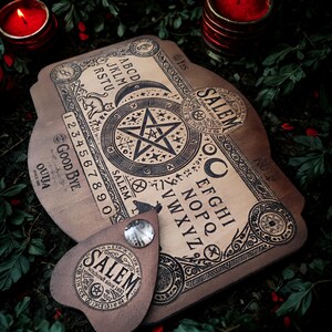 May include: A wooden Ouija board with a planchette, featuring engraved letters, numbers, and symbols. The board has the word "Salem" and the text "Witchcraft since 1692". Two red candles and greenery are in the background.