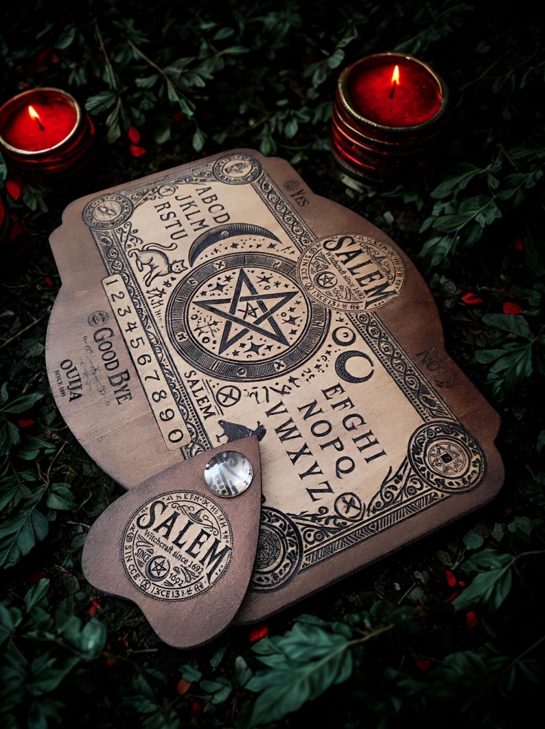 May include: A wooden Ouija board with a planchette, featuring engraved letters, numbers, and symbols. The board has the word "Salem" and the text "Witchcraft since 1692". Two red candles and greenery are in the background.