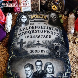 May include: A decorative Ouija board featuring the Buffy the Vampire Slayer theme. The board has a black border and a distressed, aged appearance. It includes the alphabet, numbers, and the words "Yes," "No," and "Good Bye." The board also features portraits of characters.