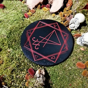 Pode incluir: Um tabuleiro circular de madeira preto com um pentagrama vermelho e outros s&iacute;mbolos inscritos nele. O tabuleiro &eacute; rodeado por musgo e outros elementos naturais.