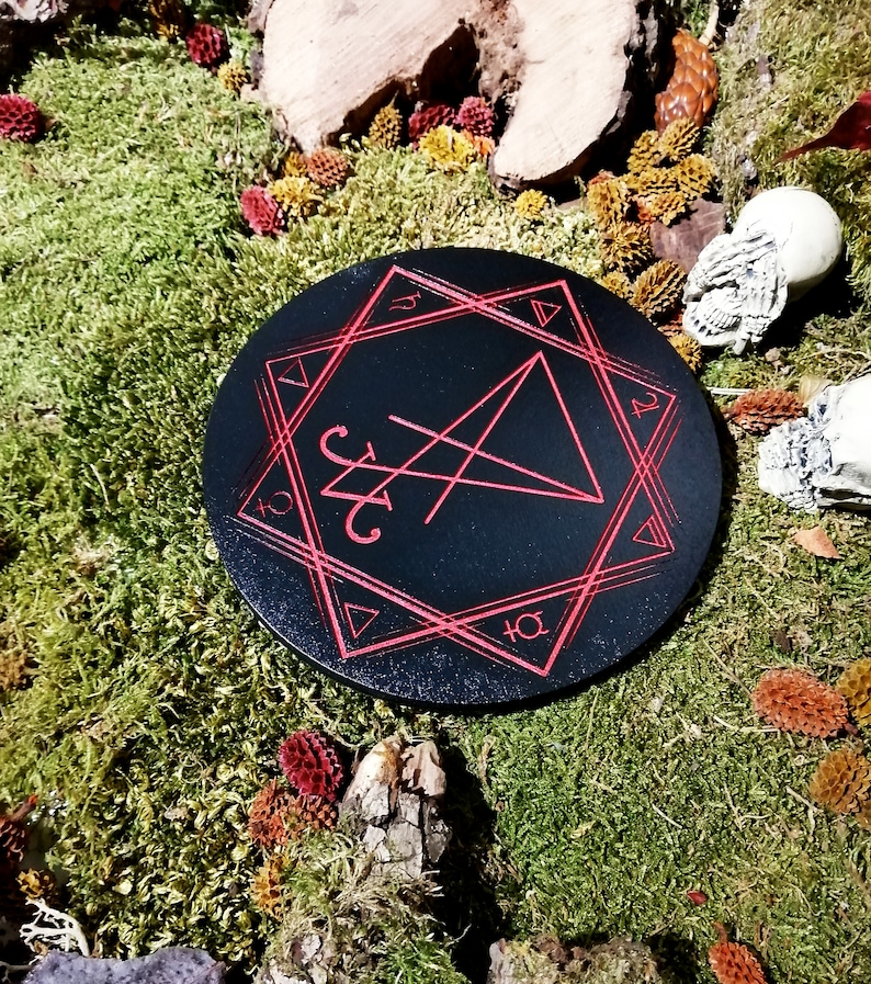 Pode incluir: Um tabuleiro circular de madeira preto com um pentagrama vermelho e outros s&iacute;mbolos inscritos nele. O tabuleiro &eacute; rodeado por musgo e outros elementos naturais.