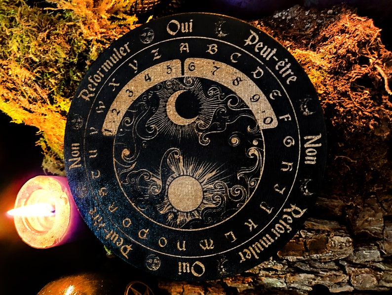 May include: A black wooden Ouija board with gold lettering and symbols. The board features a crescent moon, a sun, and swirling clouds. The text on the board reads "Oui", "Peut-&ecirc;tre", "Non", and "Reformuler".