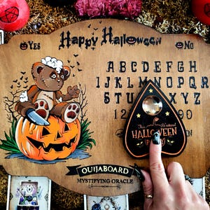 May include: Wooden Ouija board with a carved design featuring a teddy bear on a pumpkin, the words "Happy Halloween", and the alphabet. A planchette is on the right. Tarot cards and candles are in the background.
