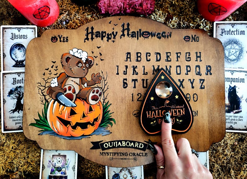May include: Wooden Ouija board with a carved design featuring a teddy bear on a pumpkin, the words "Happy Halloween", and the alphabet. A planchette is on the right. Tarot cards and candles are in the background.