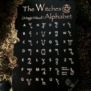 May include: Black wooden board with a gold pentagram and the text "The Witches (Magic Ritual) Alphabet". The board features a gold alphabet with symbols for each letter.