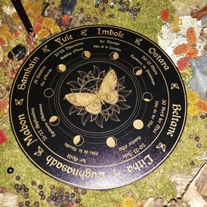 May include: A black wooden wheel with gold lettering and a gold butterfly design. The wheel is divided into eight sections, each representing a different time of year. The text on the wheel includes the names of the eight sabbats, the dates of the solstices and equinoxes, and the names of the months in French.