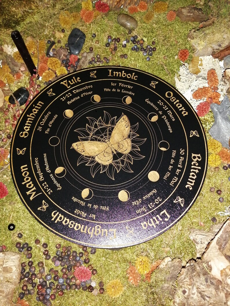 May include: A black wooden wheel with gold lettering and a gold butterfly design. The wheel is divided into eight sections, each representing a different time of year. The text on the wheel includes the names of the eight sabbats, the dates of the solstices and equinoxes, and the names of the months in French.
