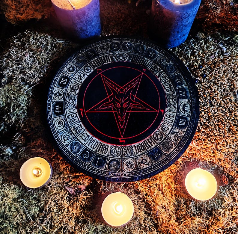 May include: A black wooden circle with a red pentagram and a goat's head inside. The circle is surrounded by a ring of symbols and numbers. The circle is on a bed of moss with three lit candles.