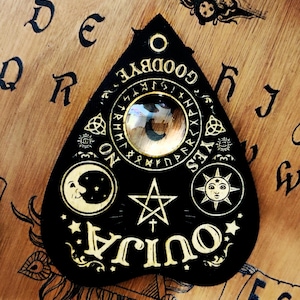 May include: Black Ouija planchette with gold lettering and symbols, including a crescent moon, sun, and star. The planchette has a clear lens in the centre. The background is a wooden surface with letters and numbers.