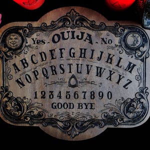 May include: A vintage-style Ouija board made of wood, featuring the alphabet, numbers, and the words "Yes," "No," and "Good Bye." The board has ornate black detailing and is surrounded by other mystical items, including a crystal ball and red objects.