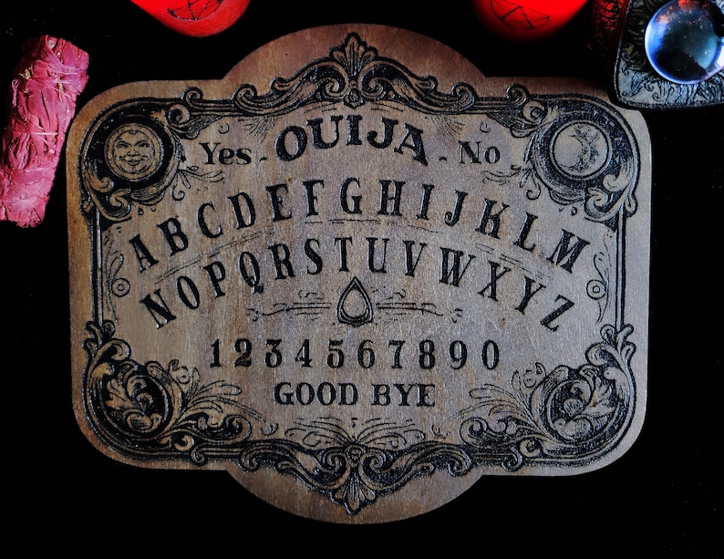May include: A vintage-style Ouija board made of wood, featuring the alphabet, numbers, and the words "Yes," "No," and "Good Bye." The board has ornate black detailing and is surrounded by other mystical items, including a crystal ball and red objects.