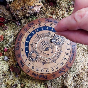 May include: A wooden Ouija board with a hand with an eye in the center. The board has a black and white design with the words "Oui", "Non", "Peut-&ecirc;tre", and "Reformuler" around the edge. The board is surrounded by moss and other natural elements.