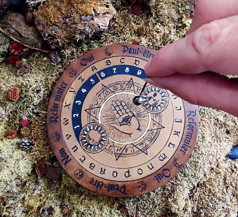 May include: A wooden Ouija board with a hand with an eye in the center. The board has a black and white design with the words "Oui", "Non", "Peut-&ecirc;tre", and "Reformuler" around the edge. The board is surrounded by moss and other natural elements.