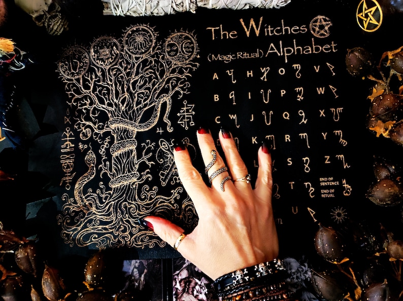 May include: A black background with a gold tree and snake design. The text "The Witches (Magic Ritual) Alphabet" is written in gold lettering. The alphabet is written in gold symbols.