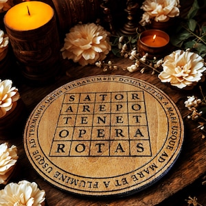 Puede incluir: Un posavasos de madera redondo con el dise&ntilde;o del Cuadrado Sator y texto. Est&aacute; sobre una superficie de madera, con velas y flores en el fondo. El dise&ntilde;o es negro sobre la madera natural. Se ven velas y flores.