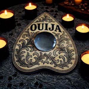 May include: A wooden Ouija board with the word "OUIJA" in black lettering, surrounded by ornate black floral designs. A glass planchette sits in the center. Lit candles in black containers surround the board, creating a mystical ambiance.