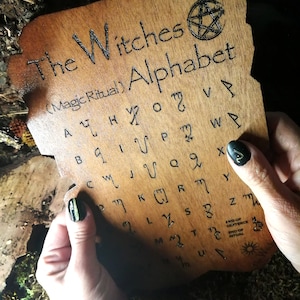 Puede incluir: Una tabla de madera con un pentagrama y el texto "The Witches (Magic Ritual) Alphabet" y una cuadr&iacute;cula de letras en una fuente &uacute;nica.