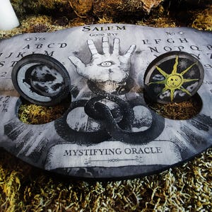 K&ouml;nnte beinhalten: Ein schwarzes Ouija-Brett aus Holz mit einer wei&szlig;en Hand mit einem Auge in der Mitte, einem schwarz-wei&szlig;en Mond und einer gelben Sonne. Das Brett hat die Buchstaben des Alphabets, die Zahlen 1-9 und die W&ouml;rter "Ja", "Nein", "Leb wohl" und "Salem".