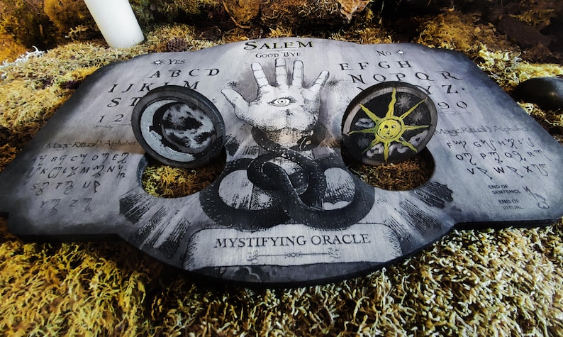 K&ouml;nnte beinhalten: Ein schwarzes Ouija-Brett aus Holz mit einer wei&szlig;en Hand mit einem Auge in der Mitte, einem schwarz-wei&szlig;en Mond und einer gelben Sonne. Das Brett hat die Buchstaben des Alphabets, die Zahlen 1-9 und die W&ouml;rter "Ja", "Nein", "Leb wohl" und "Salem".